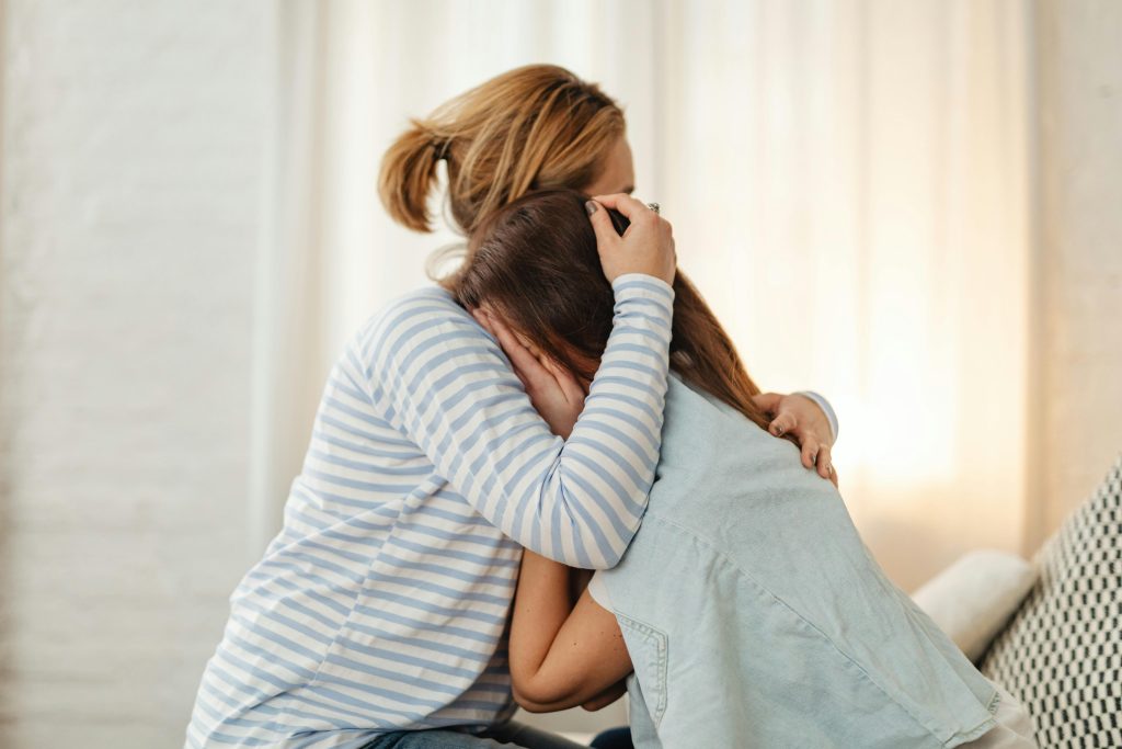 A mother lovingly comforts her teenage daughter in an emotional embrace indoors.